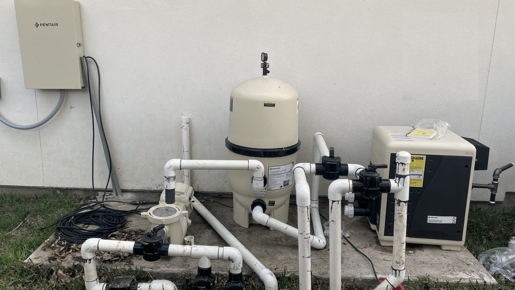 An outdoor equipment pad with a filter tank, a pump and a PVC manifold against a wall.