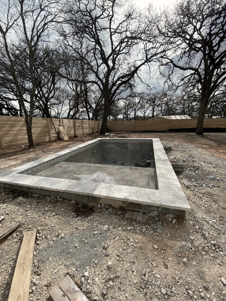 A rectangular concrete basin set in bare ground, with a wooden fence and leafless trees behind it.