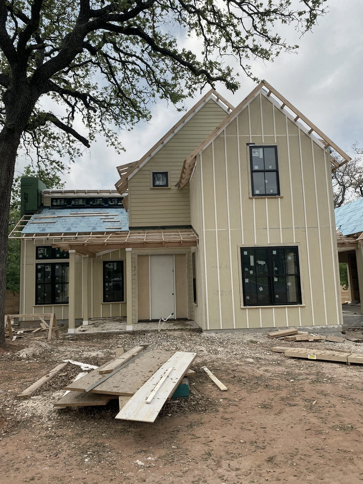 A new build with pale board-and-batten siding under a large oak, loose boards lying on the dirt in front.