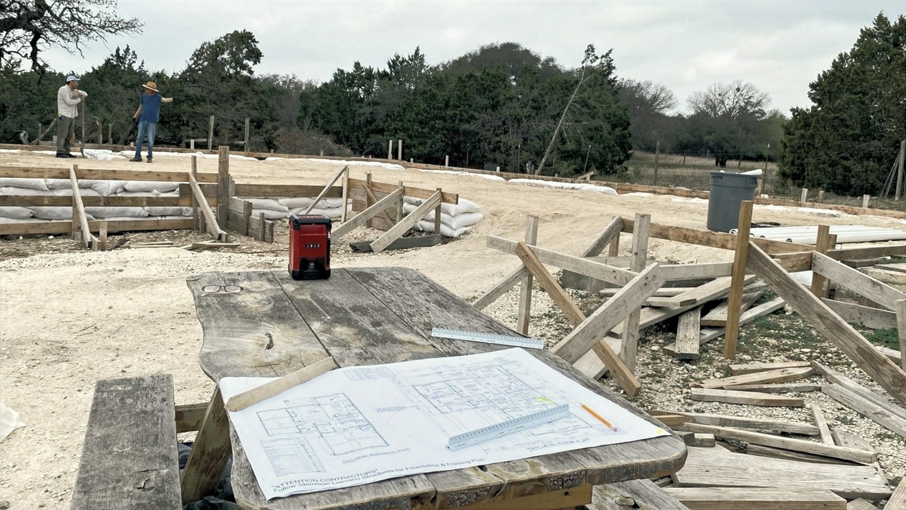 A foundation being formed with timber bracing and sandbags, and a plan sheet weighted down on a table.
